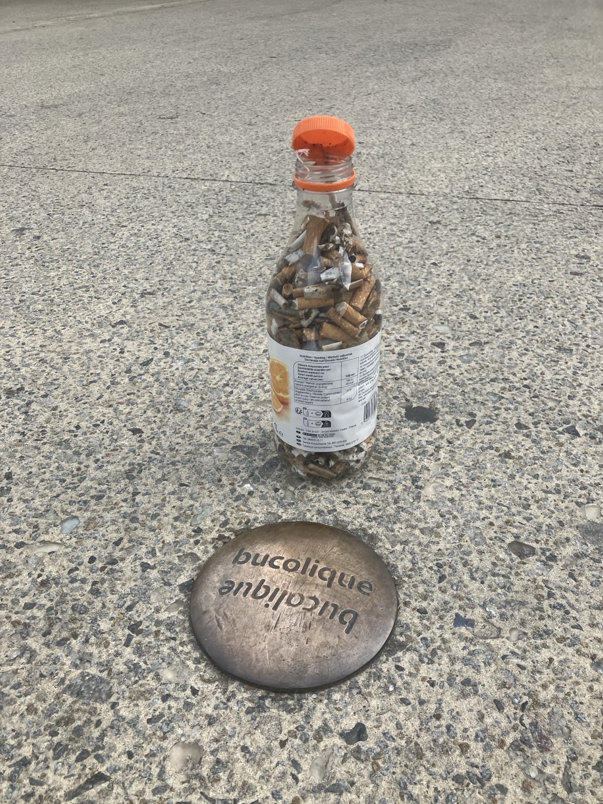 Une boouteille en plastique rempli de mégots de cigarette et au sol l'inscription "bucolique"